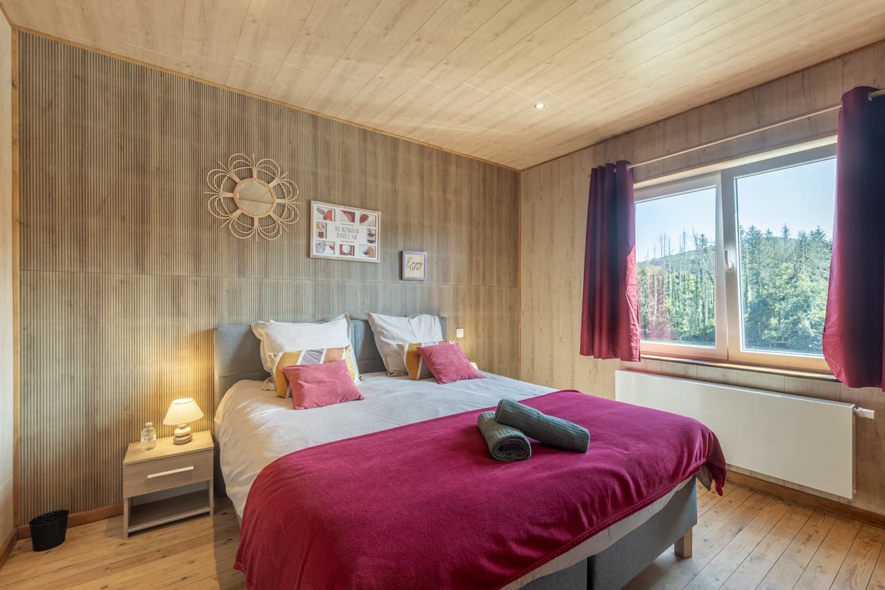 Houten slaapkamer met frambooskleurige sprei en raam op het bos
